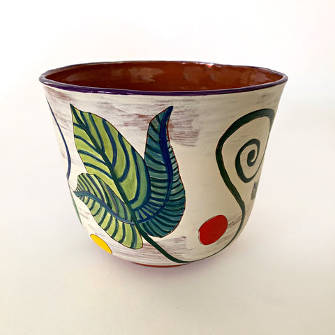 Ceramic vessel with clay red interior and white exterior decorated with bold green and blue plant leaves, with red and yellow dots surrounding, by artist Sara Schneidman at Cottage Curator - Sperryville VA Art Gallery